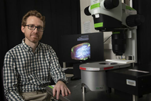 Man with red hair, beard and glasses wearing black & white checked shirt sitting next to large microscope.