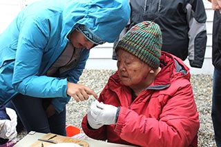 Two people in parkas, one on left with hood up blue-green, the other seated orange wearing latex gloves and looking at scientific sample together